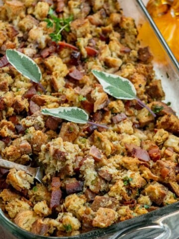 Close-up of golden, crispy sourdough stuffing with smoky kielbasa and bacon in a glass baking dish, garnished with fresh sage leaves.