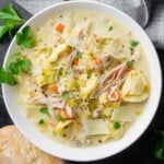 Creamy chicken tortellini soup with leeks, shredded chicken, carrots, Parmesan, and parsley in a white bowl.