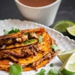 Crock pot Birria Tacos with cilantro, onion, and lime with a side of cxonsommé.