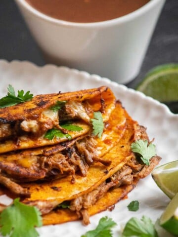 Crock pot Birria Tacos with cilantro, onion, and lime with a side of cxonsommé.