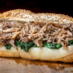 Close-up of a South Philly Italian roast pork sandwich stuffed with tender shredded pork, garlicky spinach, and melted provolone cheese on a seeded hoagie roll.