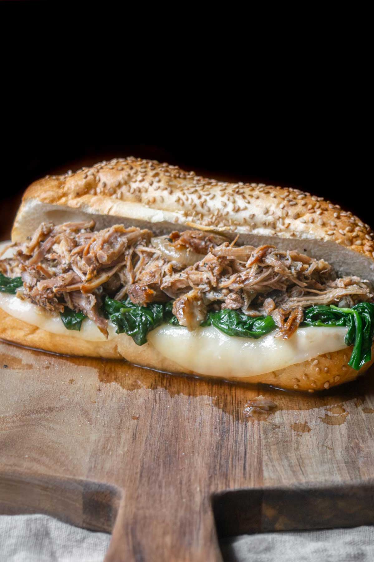 South Philly Italian roast pork sandwich piled high with tender shredded pork, melted provolone, and garlicky spinach on a seeded Italian roll, served on a wooden cutting board.