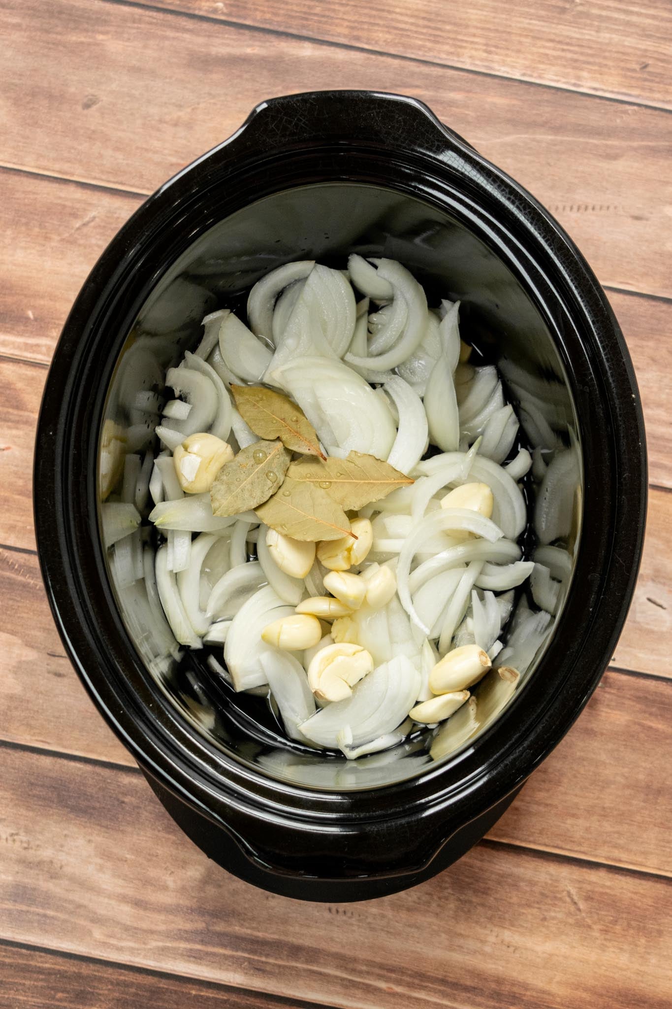 Get your slow cooker ready. Add the sliced onions, peeled garlic cloves, wine, and bay leaves to the bottom of your crockpot. I recommend a 7-quart slow cooker for the full 8-pound pork shoulder.