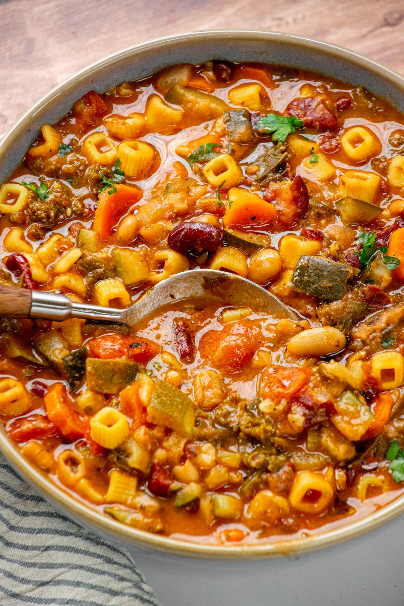 Spoon resting in a hearty bowl of vegetarian minestrone soup packed with pasta, beans, zucchini, carrots, and fresh parsley.