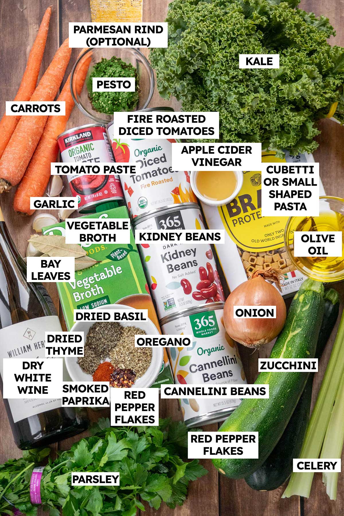 Overhead view of labeled vegetarian minestrone soup ingredients, including zucchini, kale, carrots, beans, cubetti pasta, fire-roasted tomatoes, pesto, vegetable broth, herbs, and spices.