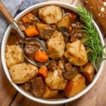 Close-up of slow cooker beef stew and dumplings with tender beef, fluffy biscuit dumplings, red potatoes, carrots, and mushrooms in rich gravy, garnished with rosemary.