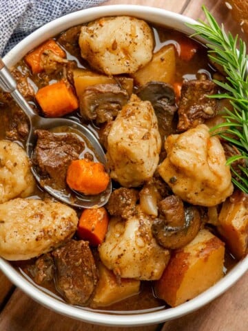 Close-up of slow cooker beef stew and dumplings with tender beef, fluffy biscuit dumplings, red potatoes, carrots, and mushrooms in rich gravy, garnished with rosemary.