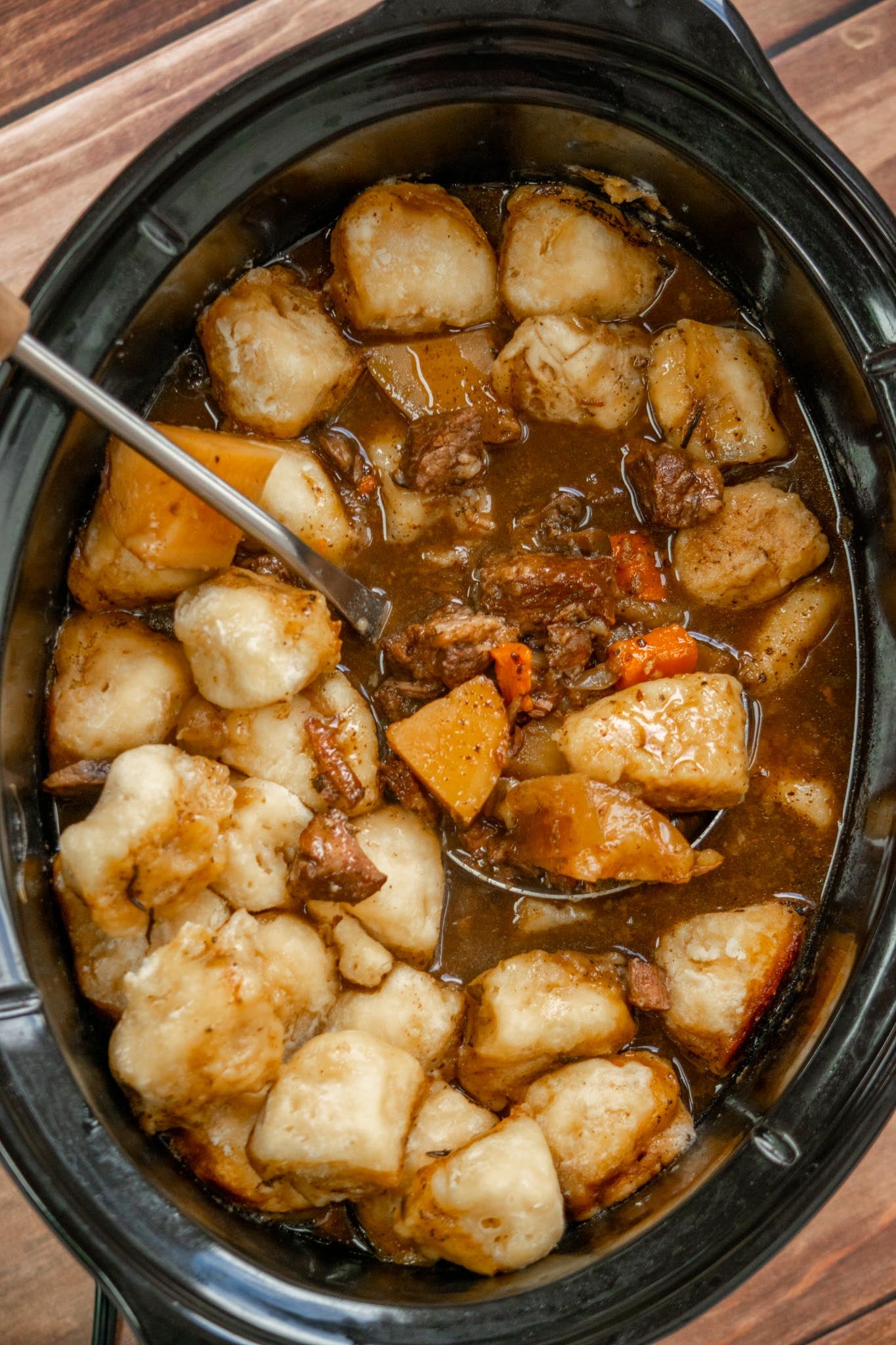 Finished crockpot beef stew with biscuit dumplings, tender beef, carrots, and potatoes in a rich brown gravy, with a serving spoon in the slow cooker.