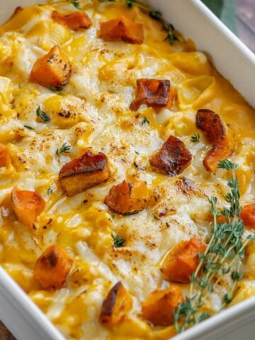 Creamy baked butternut squash mac and cheese topped with golden roasted squash cubes, melted cheddar cheese, and fresh thyme in a white baking dish.