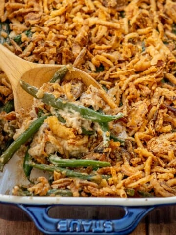 Homemade green bean casserole with fresh green beans in a creamy mushroom sauce, topped with crispy fried onions and served with a wooden spoon in a blue baking dish.