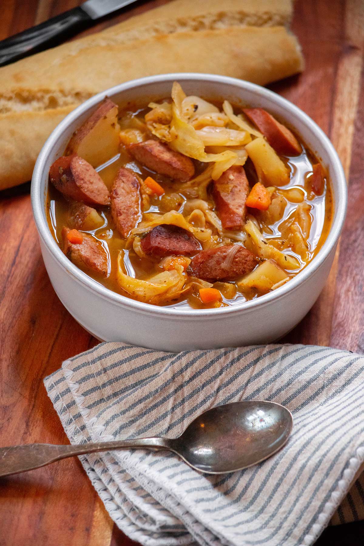 Bowl of cabbage soup with sliced kielbasa, red potatoes, carrots and cabbage served with crusty bread.