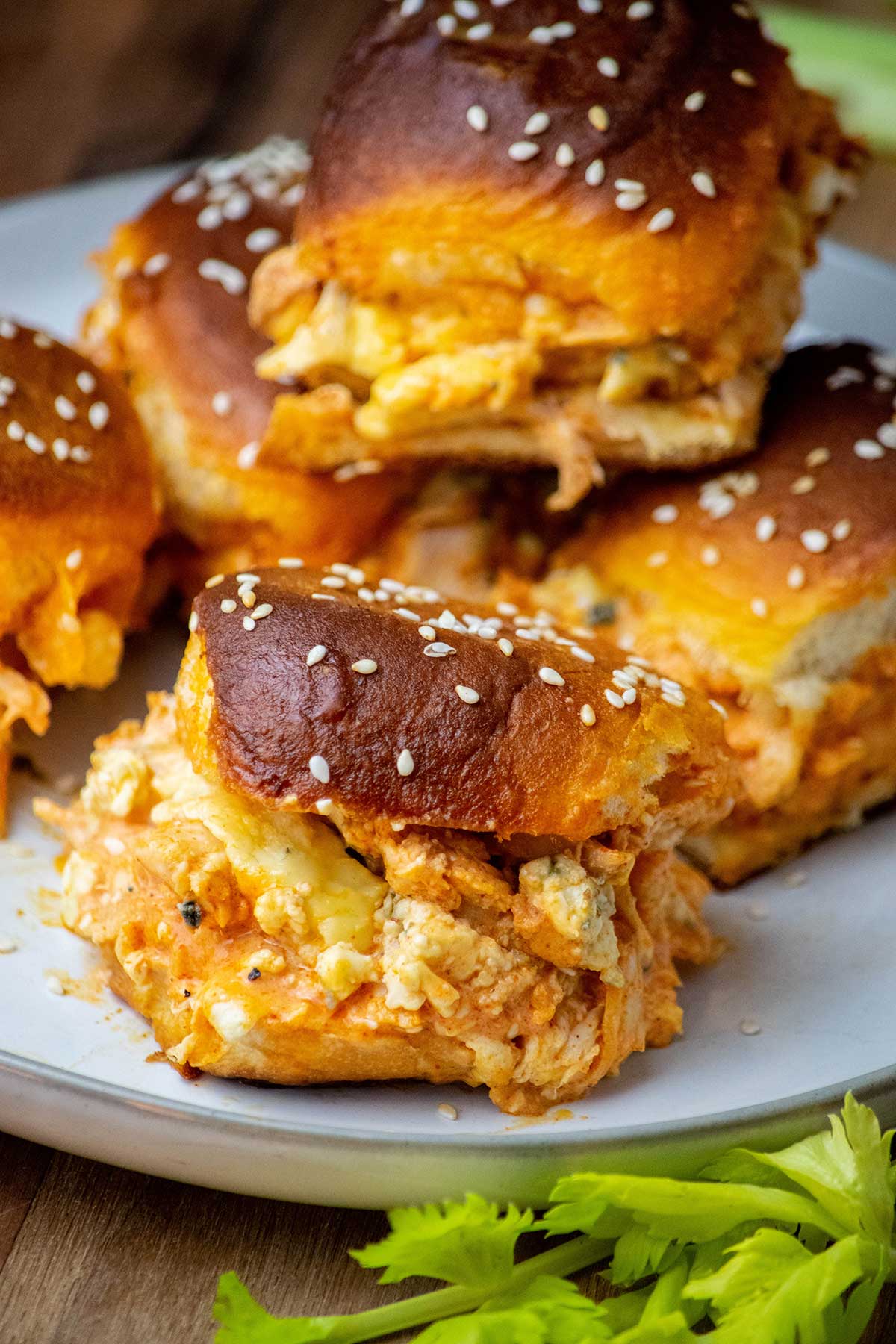 Plate of Buffalo chicken sliders with spicy chicken, melted cheese, blue cheese crumbles, and sesame-topped rolls, served with celery.