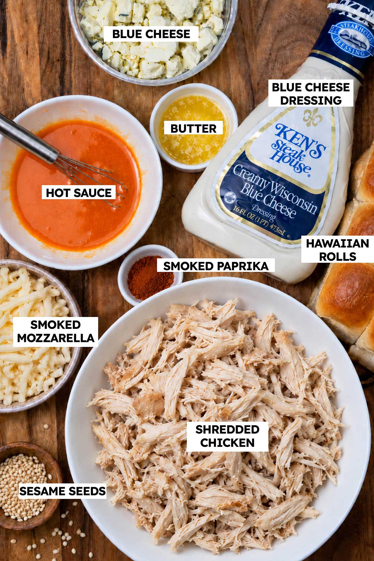 Ingredients for Buffalo Chicken Sliders, including shredded chicken, buffalo sauce, blue cheese dressing, smoked mozzarella, blue cheese crumbles, Hawaiian rolls, butter, sesame seeds, and smoked paprika.