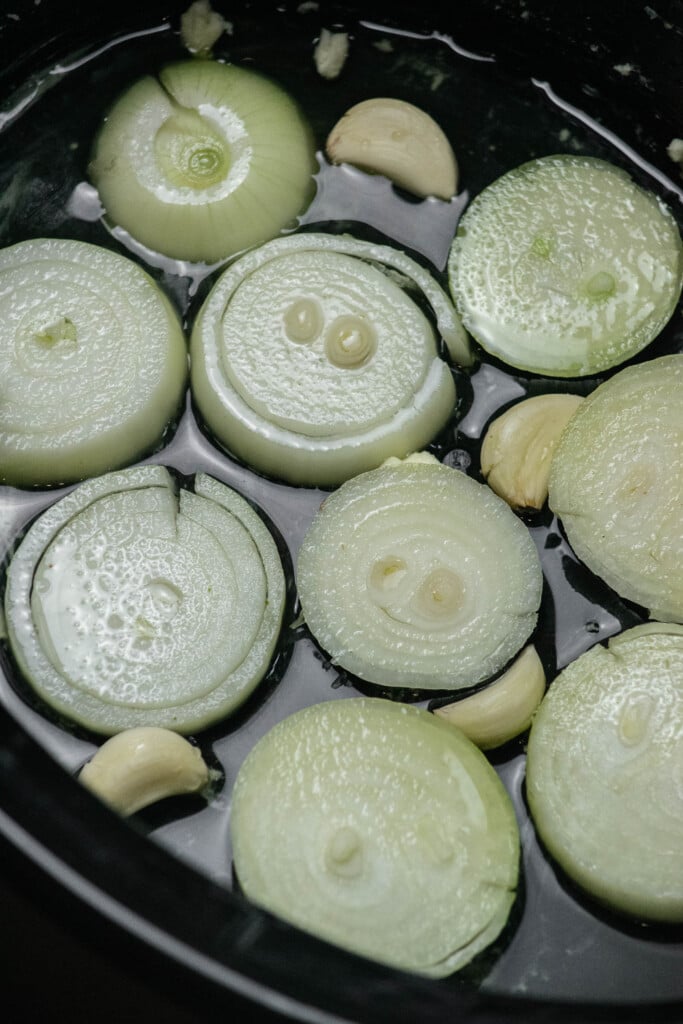 Butter the crockpot liberally to prevent sticking, and pour in chicken stock. Shingle onion slices in the bottom, and add the garlic cloves.