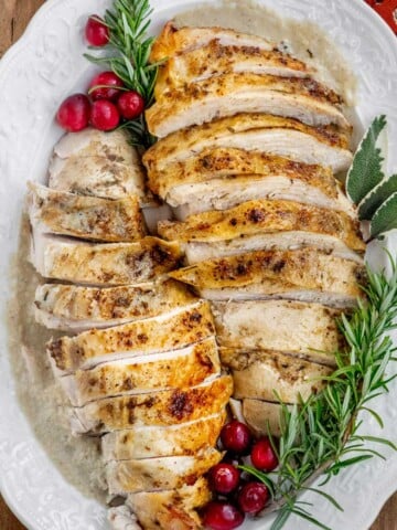 Sliced Crockpot turkey breast with golden herb-seasoned skin and homemade onion gravy, garnished with fresh rosemary, sage, and cranberries.