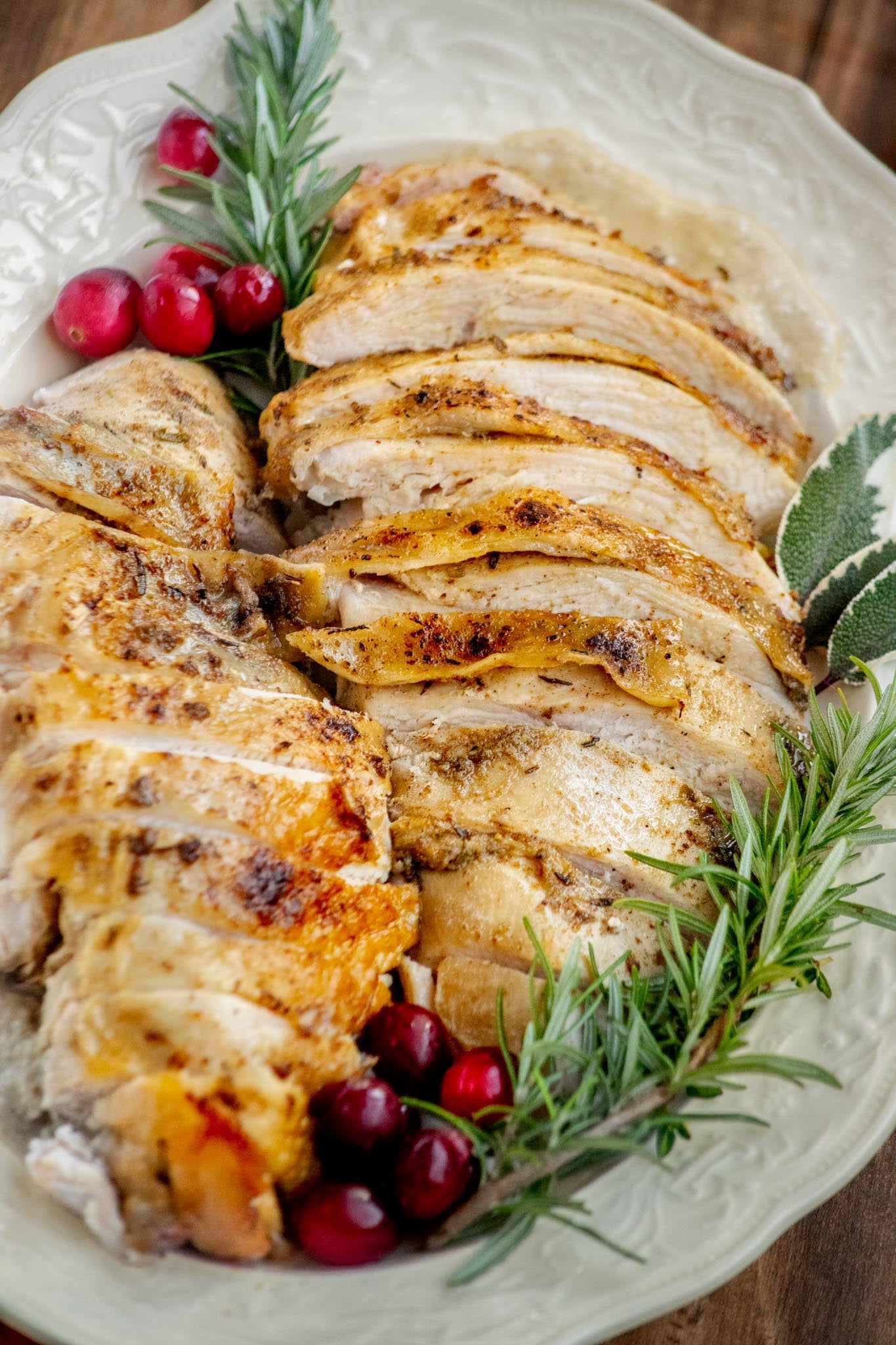 Sliced Crockpot turkey breast with golden herb-seasoned skin and homemade onion gravy, garnished with fresh rosemary, sage, and cranberries on a white platter.