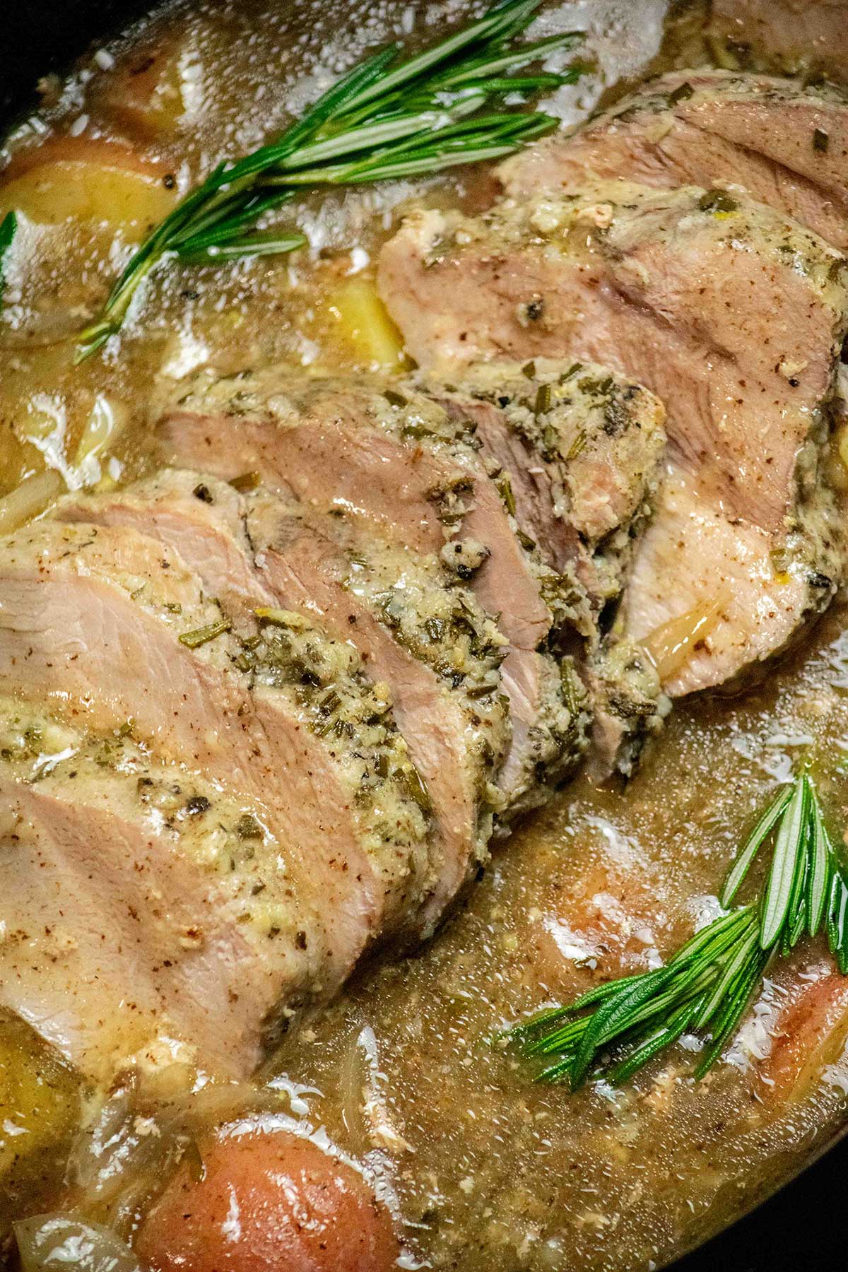Close-up of sliced slow cooker pork sirloin roast with gravy, red potatoes, onions, and fresh rosemary.