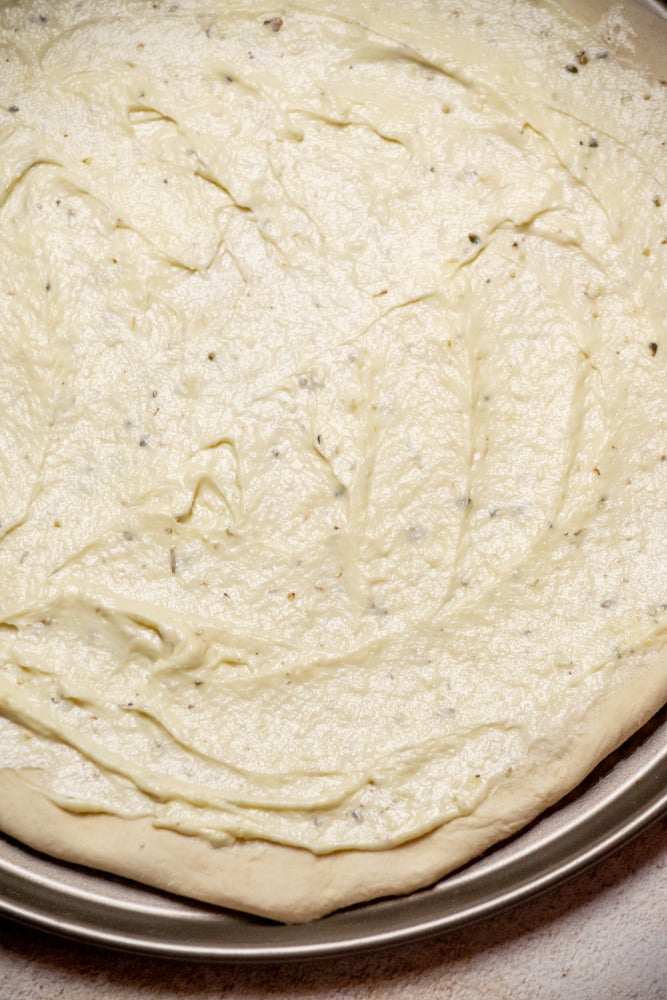 Stretch your dough to fit your pizza pan, and spread the cooled white sauce over the dough, leaving about ¼" gap at the edges.