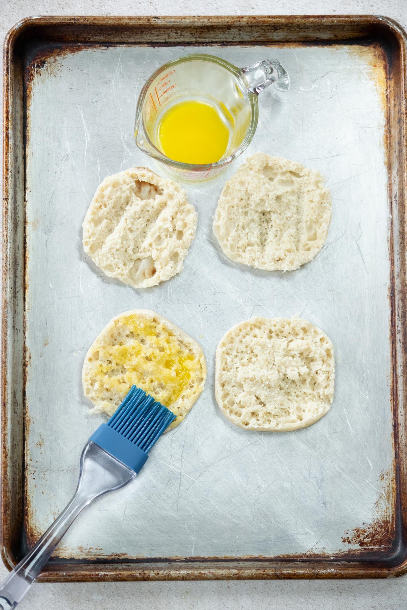 English muffins split on a baking tray with a pastry brush adn dish of melted butter.