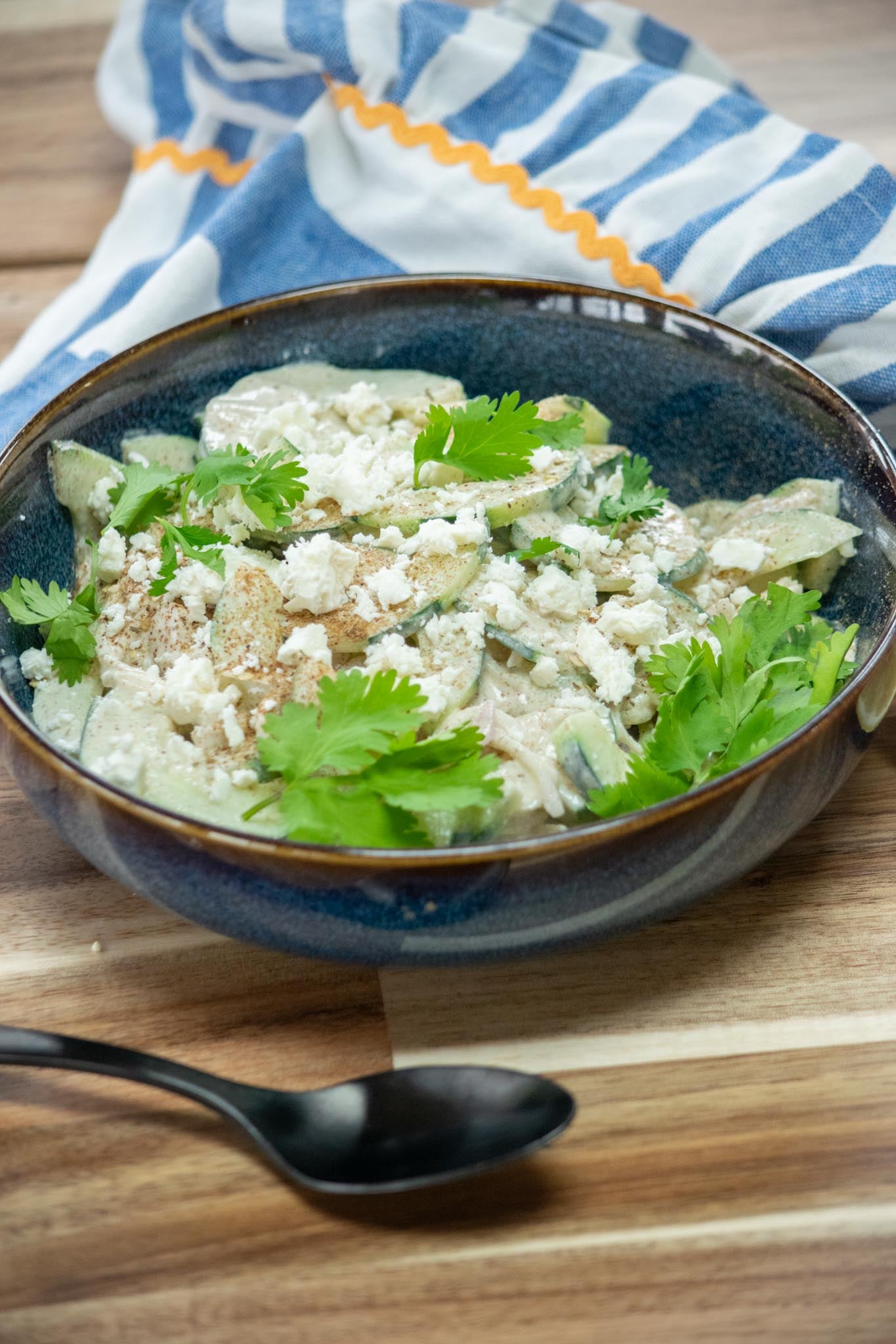 A bowl of cucumber salad with feta in front of a blue and white napkin.