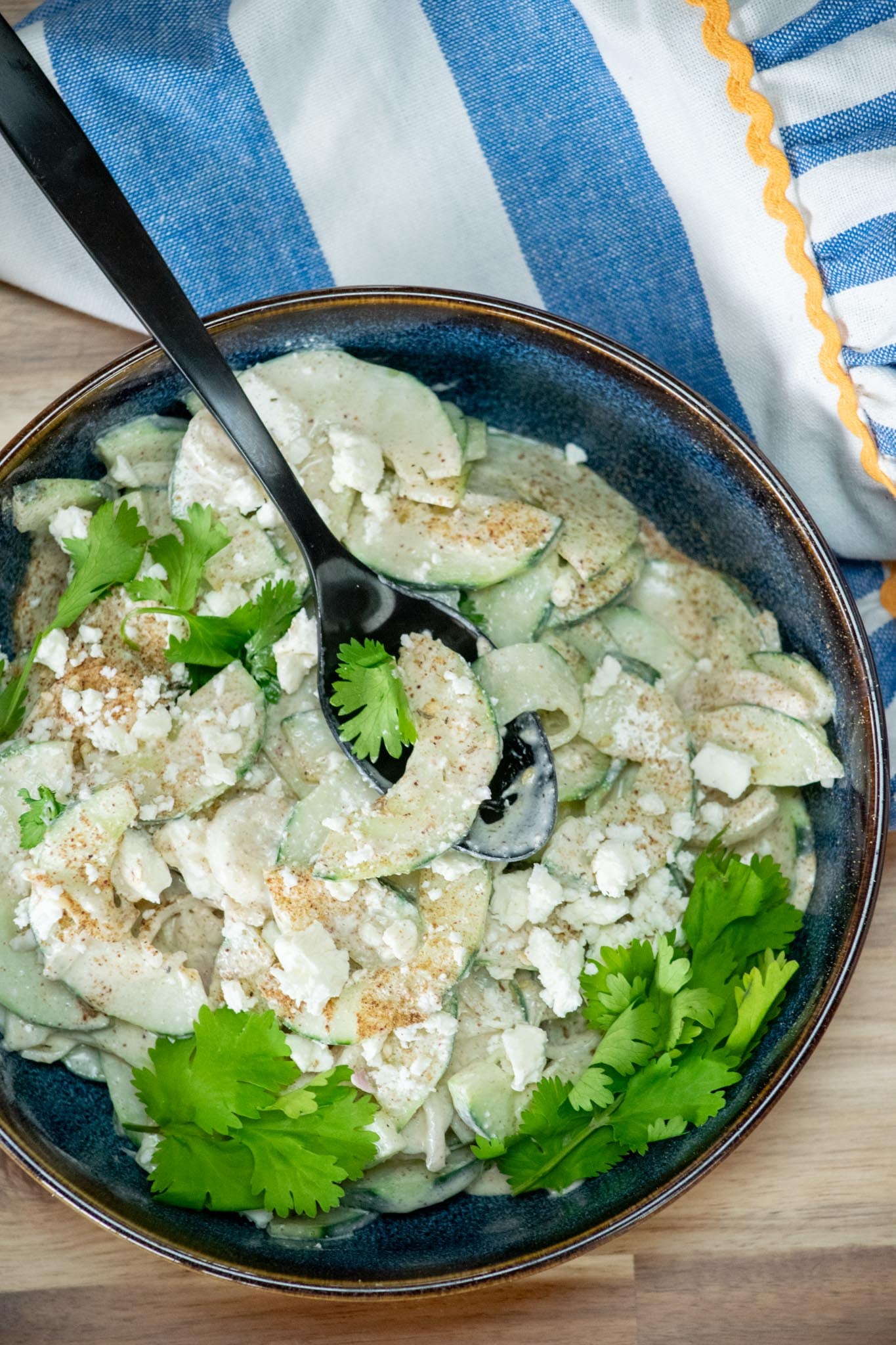 A bowl of cucumber salad topped with feta cheese crumbles and a spoon in the bowl.