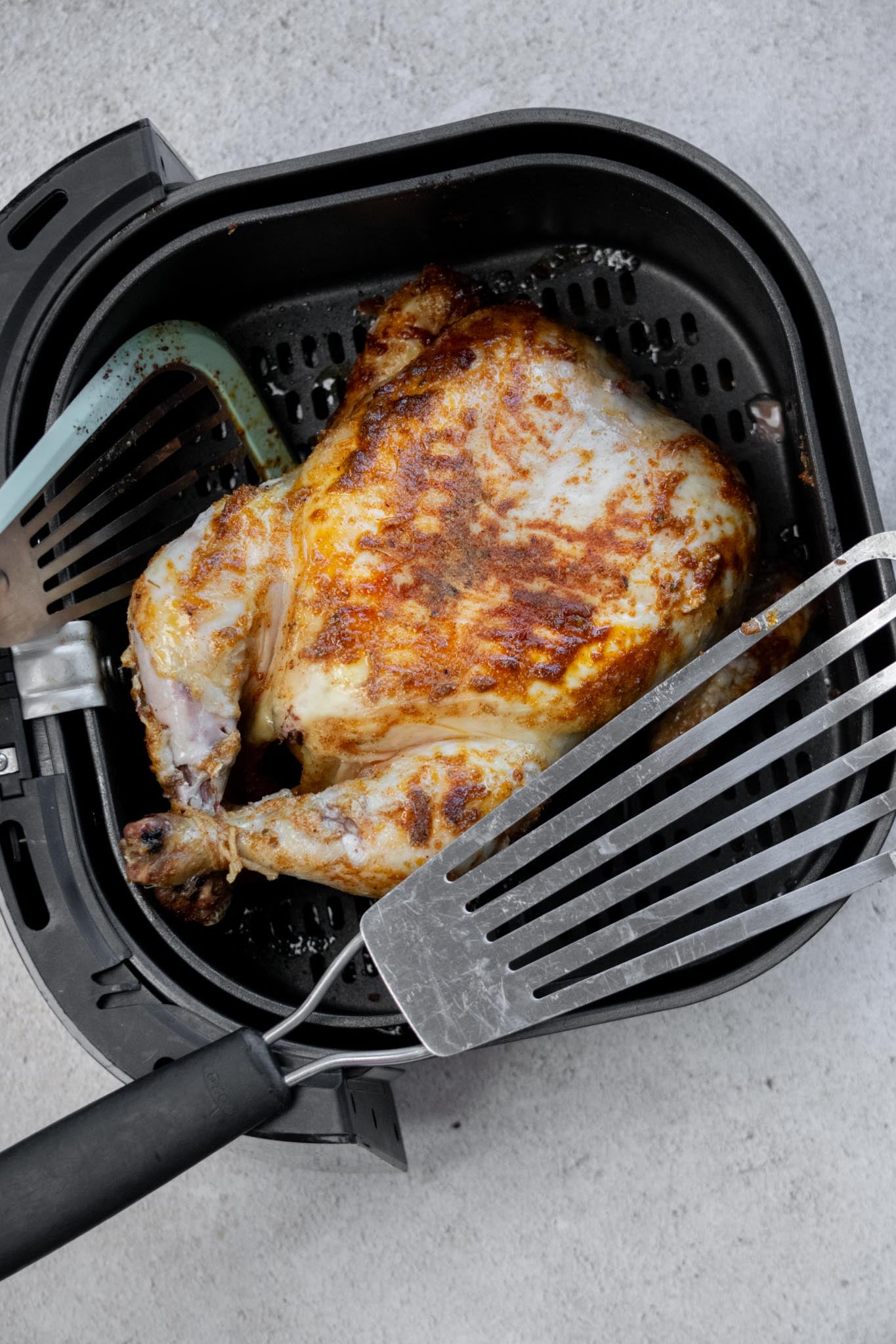 Whole roasted chicken in the air fryer basket with a spatula to the side.